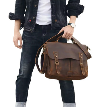 About Us Heritage Legacy Carry in brown genuine crazy horse leather with distressed finish held by hand featuring top handle front buckle closures removable adjustable shoulder strap and vintage silhouette showcasing rugged elegance and upscale everyday styling