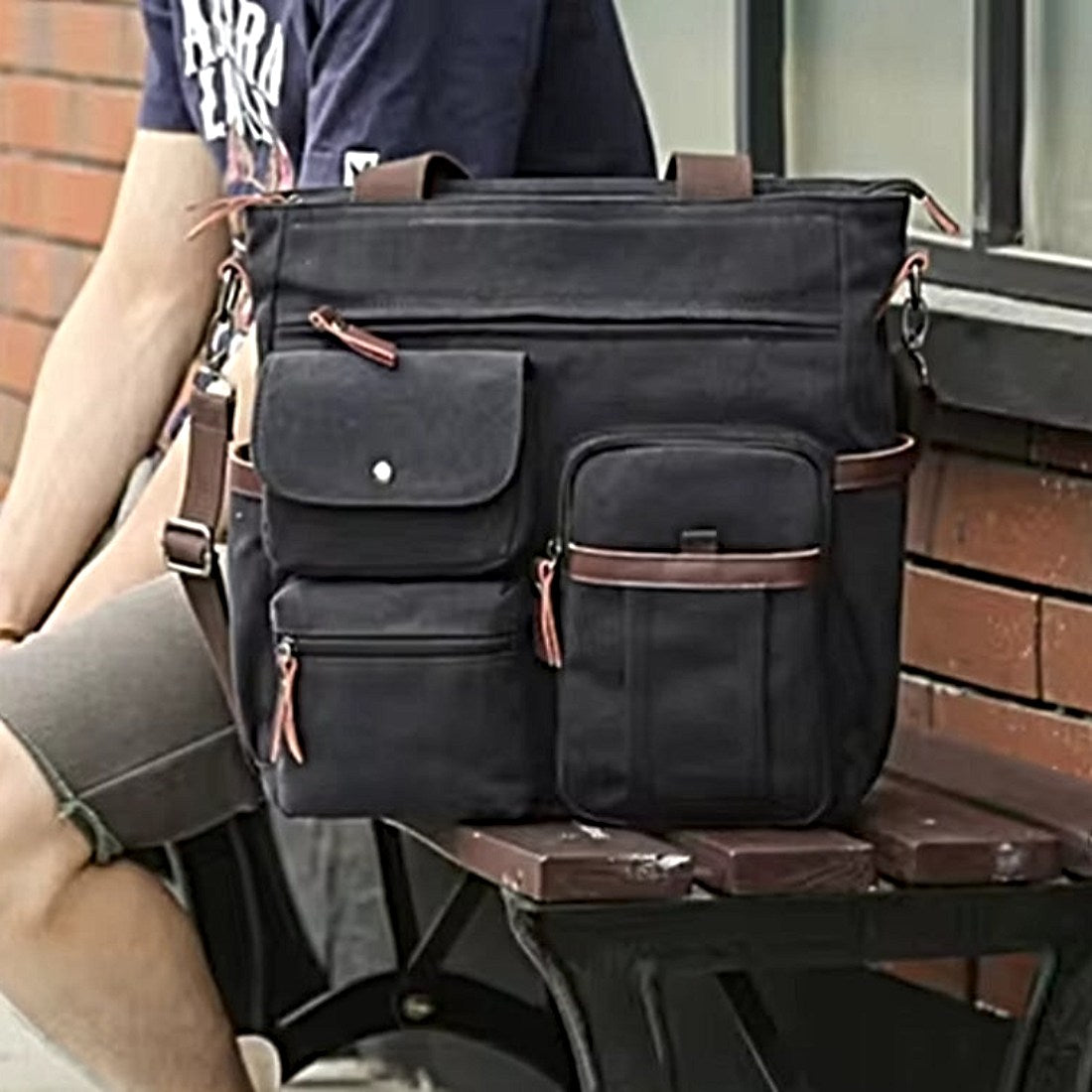 Nomad Utility Carry bag in black durable canvas with brown leather accents shown resting on bench beside model featuring multiple compartments zippered and flap pockets dual handles and adjustable shoulder strap showcasing durable premium construction and rugged upscale styling for life on the move