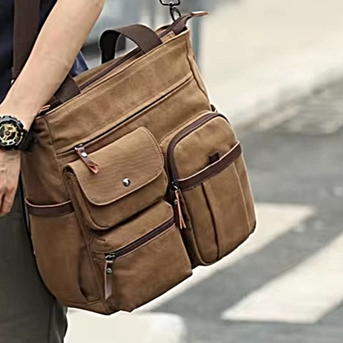 Nomad Utility Carry bag in brown durable canvas with leather accents shown held by model featuring multiple compartments zippered and flap pockets dual handles and adjustable detachable shoulder strap showcasing durable premium construction and rugged upscale styling for life on the move