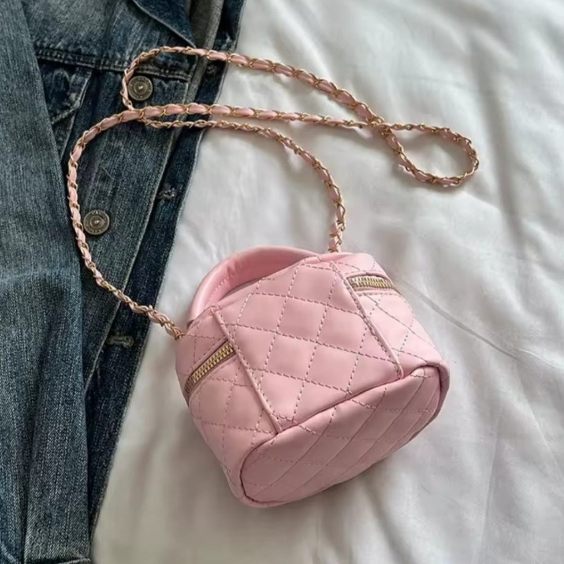 Timeless Petite Luxe elegant crossbody bag in pink quilted luxury vegan leather with upscale gold accents cylindrical silhouette with short top handle and upscale gold chain strap placed beside blue denim jeans on white surface showcasing premium design in lifestyle setting