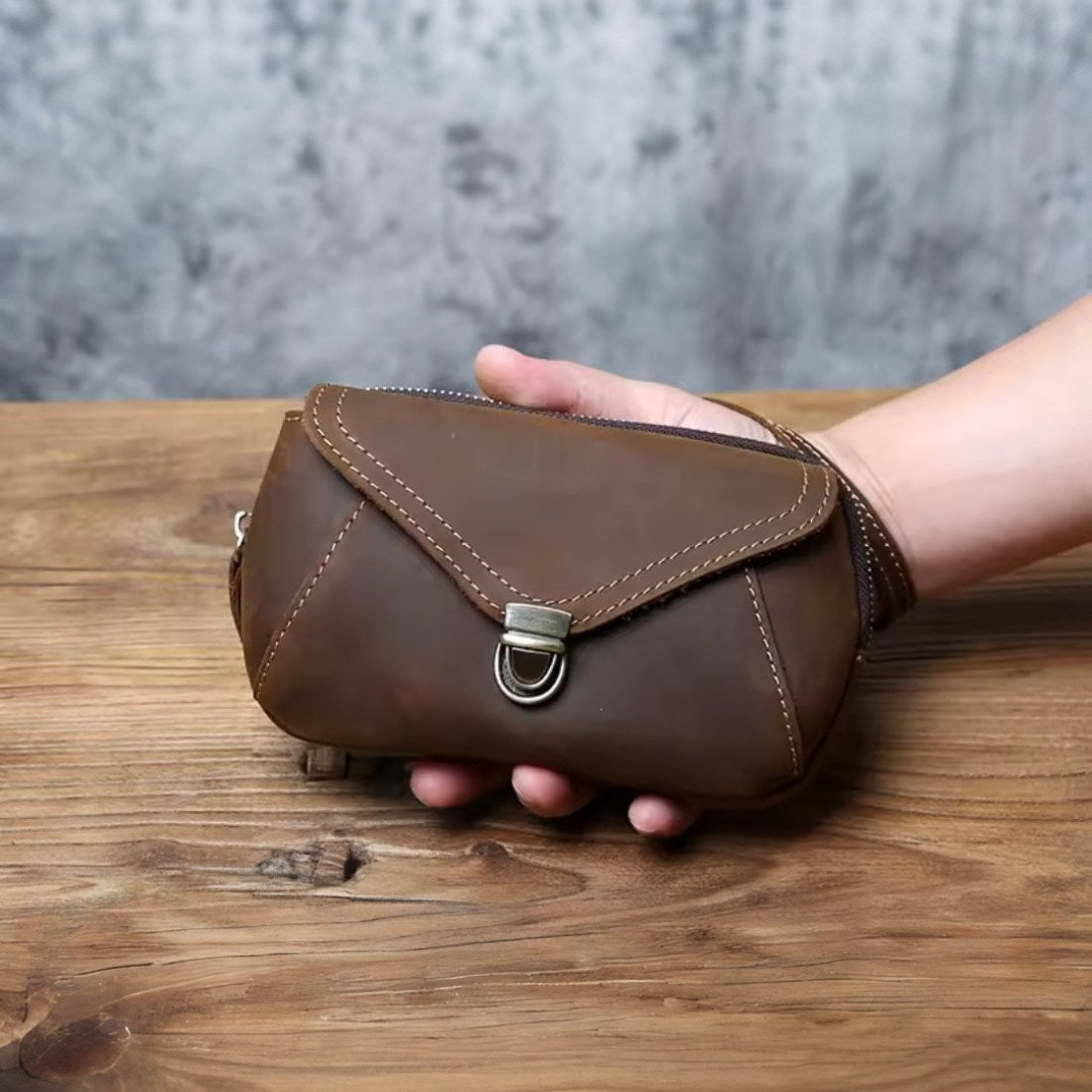 Heritage Utility Carry in coffee crazy horse genuine leather with distressed oil wax finish compact structured silhouette with triangular flap visible stitching and antique brass clasp held by hand showcasing vintage craftsmanship and upscale utility