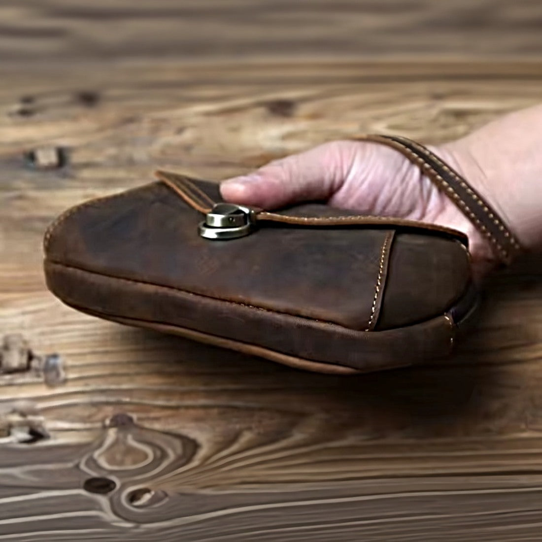Heritage Utility Carry in dark brown crazy horse genuine leather with distressed oil wax finish compact structured silhouette with visible stitching zipper closure and wrist strap held by hand over wooden surface showcasing the bottom profile highlighting vintage craftsmanship and upscale utility